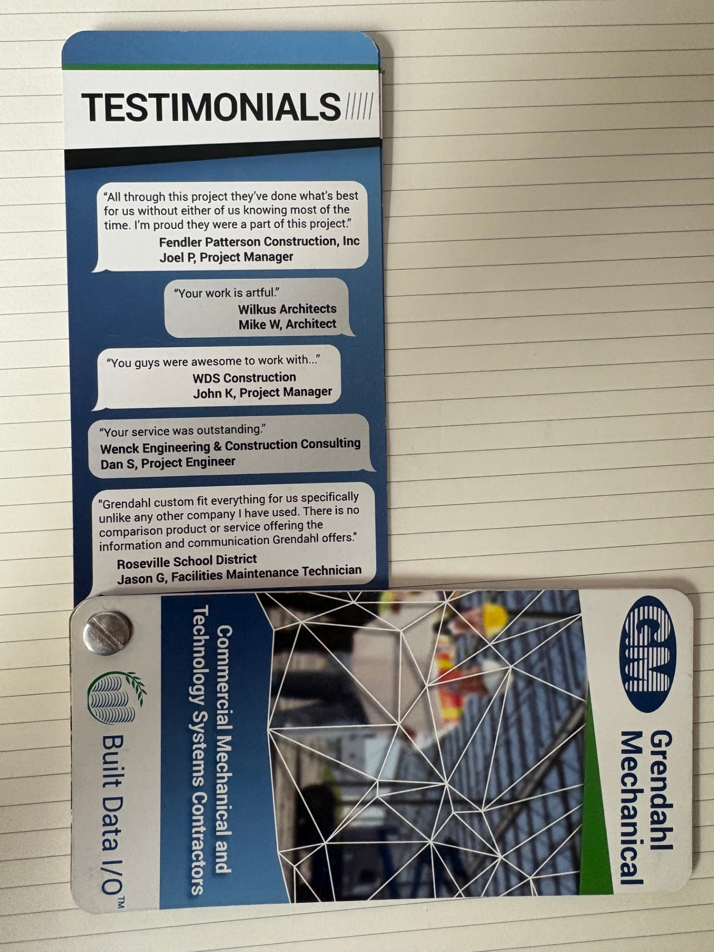 Photograph of a printed Grendahl Mechanical marketing card lying on a desk. The upper panel is headed “TESTIMONIALS” and lists five short client quotes with company names — Fendler Patterson Construction, Wilkus Architects, WDS Construction, Wenck Engineering & Construction Consulting, and Roseville School District. Below it, a folded tab shows the Grendahl Mechanical logo and vertical text reading “Built Data I/O™ — Commercial Mechanical and Technology Systems Contractors.”